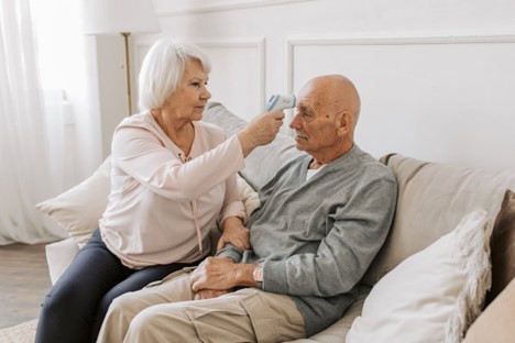 elderly woman taking temperature of eldery man at home on sofa