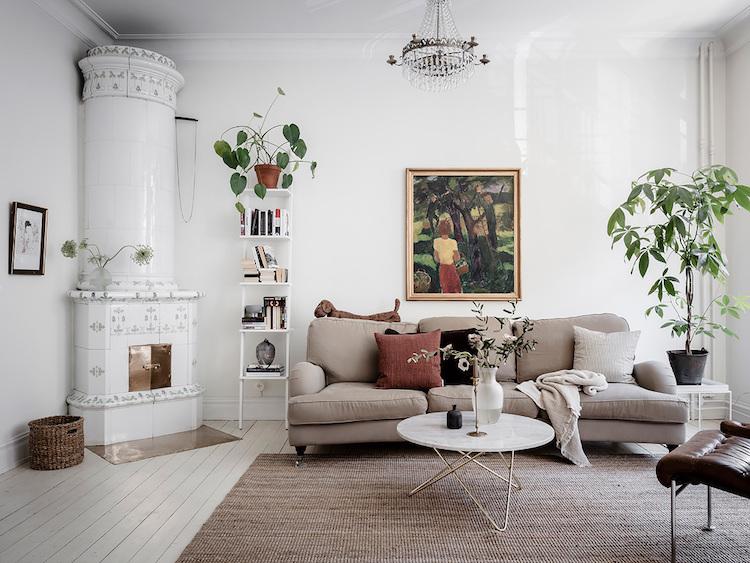 A cozy, modern living room with light gray walls, a beige sofa adorned with cushions, a white circular coffee table, and houseplants. The room features a traditional ceramic fireplace in the corner, a chandelier hanging from the ceiling, and a framed painting on the wall. A natural jute rug complements the wooden floor, creating a warm and inviting atmosphere.